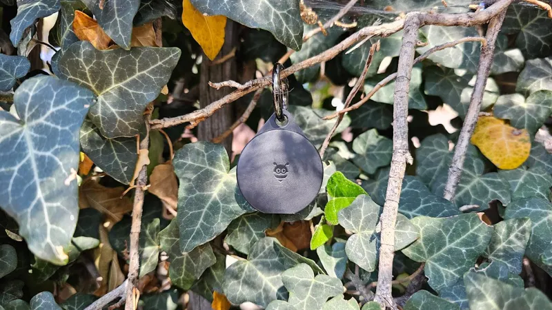 Ассортимент Bluetooth-трекеров, расположенных в виде сетки на деревянном фоне. Трекеры включают черные трекеры Tile различных форм, два серебристо-белых трекера AirTag и один круглый синий трекер Chipolo, прикрепленный к связке ключей с мультитулом.