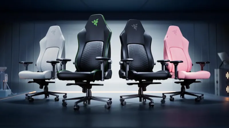 Four Razer gaming chairs — in light gray, black with green trim, black, and light pink — are pictured beside each other spot-lit in a dark room