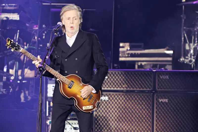 Paul McCartney playing bass live. He glances over to his side with partially pursed lips.